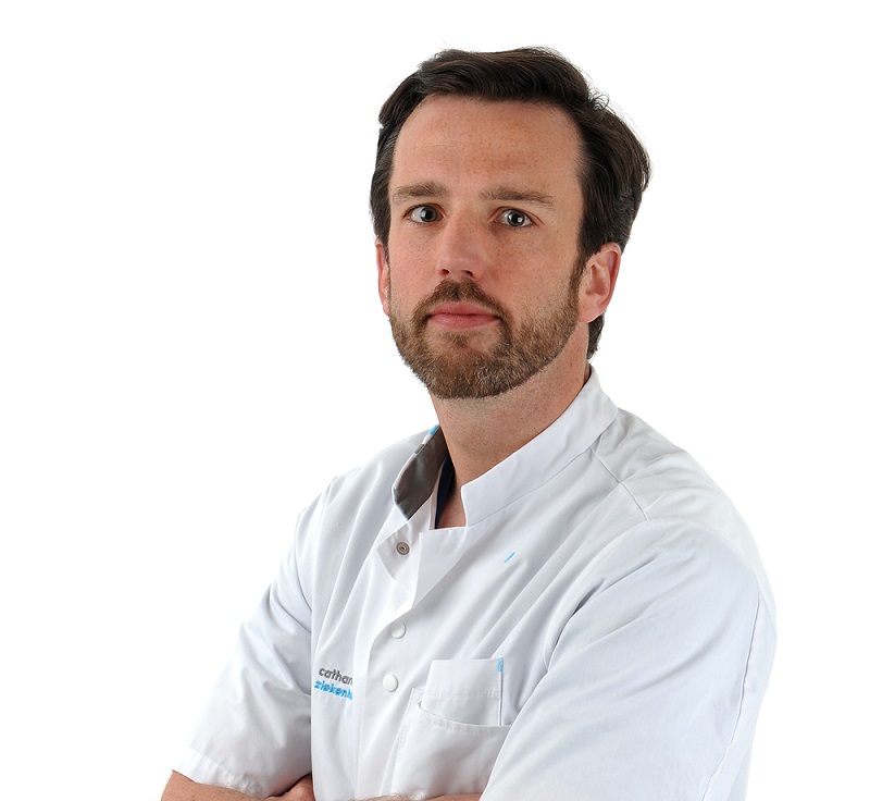 Een man met kort bruin haar en baard in een witte labjas staat met gekruiste armen voor een witte achtergrond.
