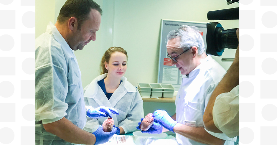 Drie mensen in labjassen en handschoenen onderzoeken biologisch materiaal in een medisch lab, met een whiteboard en labbenodigdheden.