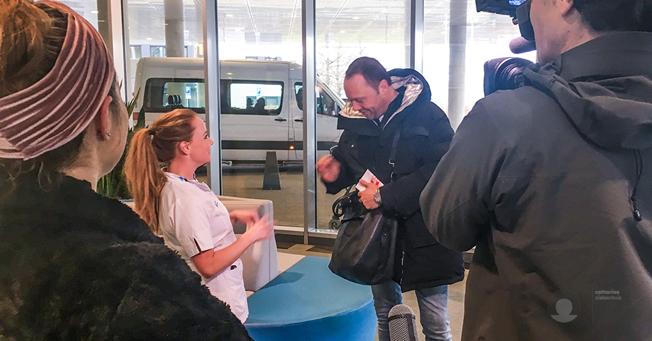 Man in winterjas en vrouw in uniform praten binnen, cameraman filmt. Andere vrouw kijkt toe. Buiten staat een busje geparkeerd.