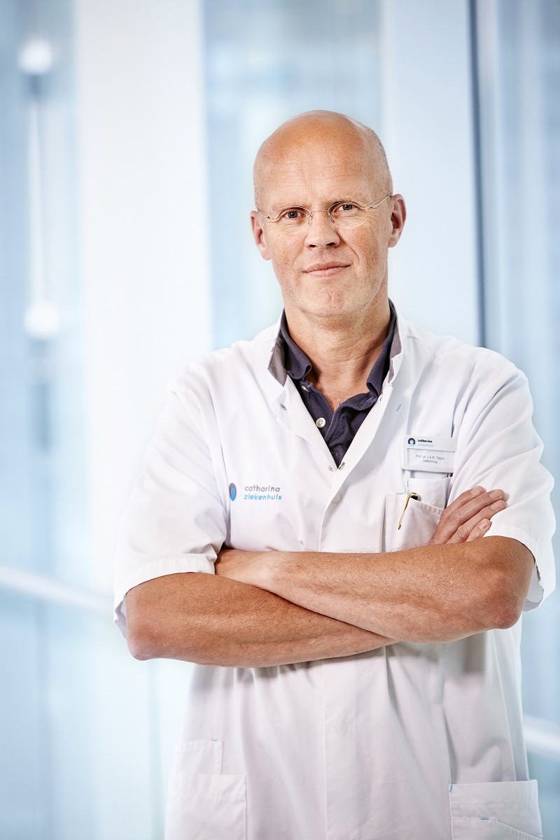Een boerenkool, middenjarige persoon met bril en neutrale blik, in witte labjas, staat met armen over elkaar in een lichte ruimte.