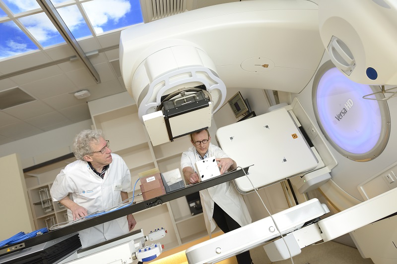Twee wetenschappers in witte jassen stellen apparatuur af onder een groot bestralingsapparaat in een heldere, kalme laboratoriumruimte.