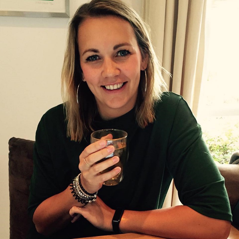 Vrouw met blond haar en groene trui zit aan tafel, glimlacht en houdt glas vast. Ze draagt sieraden en een blauwe pluis voor darmkanker.