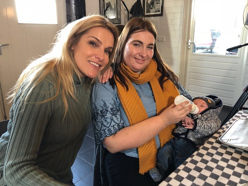 Twee vrouwen zitten aan tafel; één lacht naar de camera, de ander geeft een baby de fles. Keuken met geruite tafelkleed.