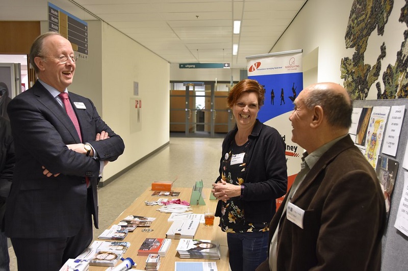 Drie volwassenen bij een Nierstichting-tafel met folders; twee praten en lachen, de ander luistert. Posters over kwaliteit van leven.