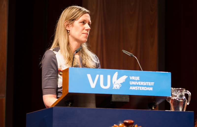 Vrouw bij spreekgestoelte met blauw VU-bord, microfoon, waterkan en glas. Houten wand op de achtergrond.