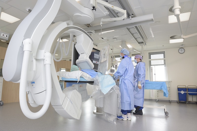 Drie medici in scrubs en maskers bedienen beeldapparatuur naast een patiënt op een ziekenhuisbed in een moderne behandelkamer.