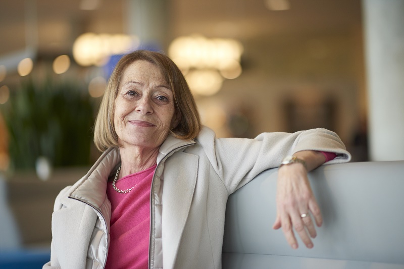 Vrouw met kort lichtbruin haar in beige jas en roze top zit op grijze bank in ziekenhuis, glimlacht naar camera.
