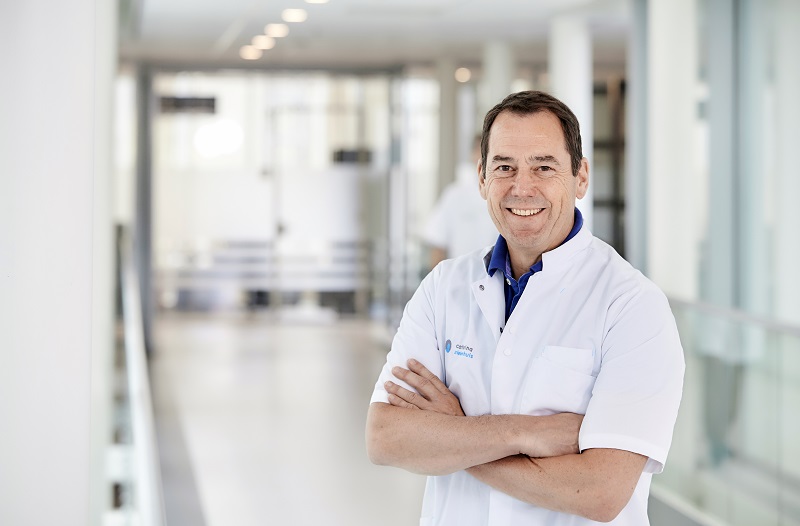 Man in witte doktersjas staat met gekruiste armen in moderne gang, lachend; lichte, ruime achtergrond is onscherp.