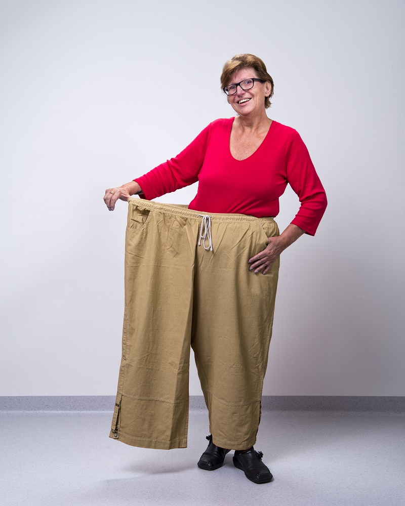 Vrouw met rode top en zwarte schoenen laat lachend zien hoe ruim een grote beige broek haar zit.