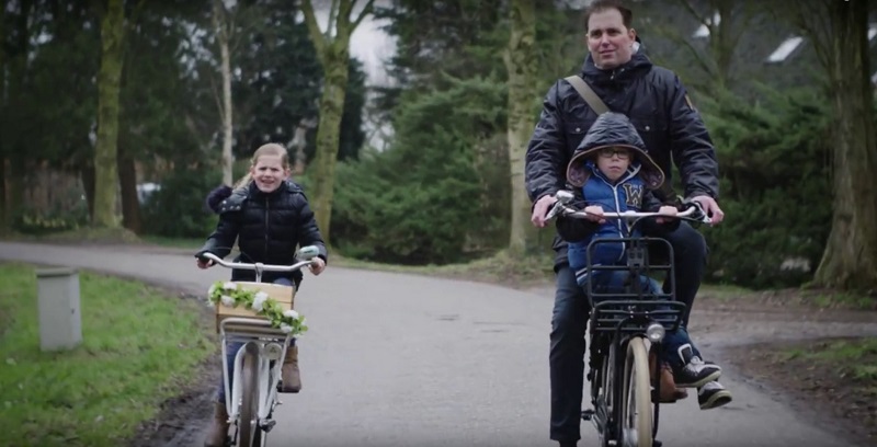 Een man en twee kinderen fietsen op een parkpad. De man en jongen delen een bakfiets, het meisje rijdt op een versierde fiets.