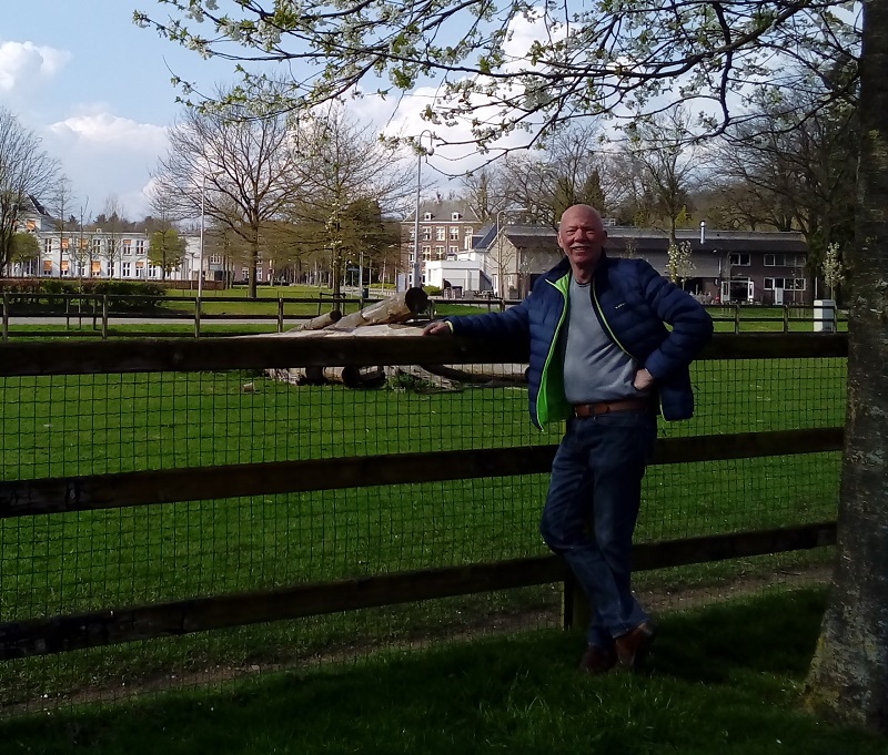 Man in blauwe jas en spijkerbroek, herstellend van trombose in arm, leunt tegen houten hek in groen park met huizen op achtergrond.