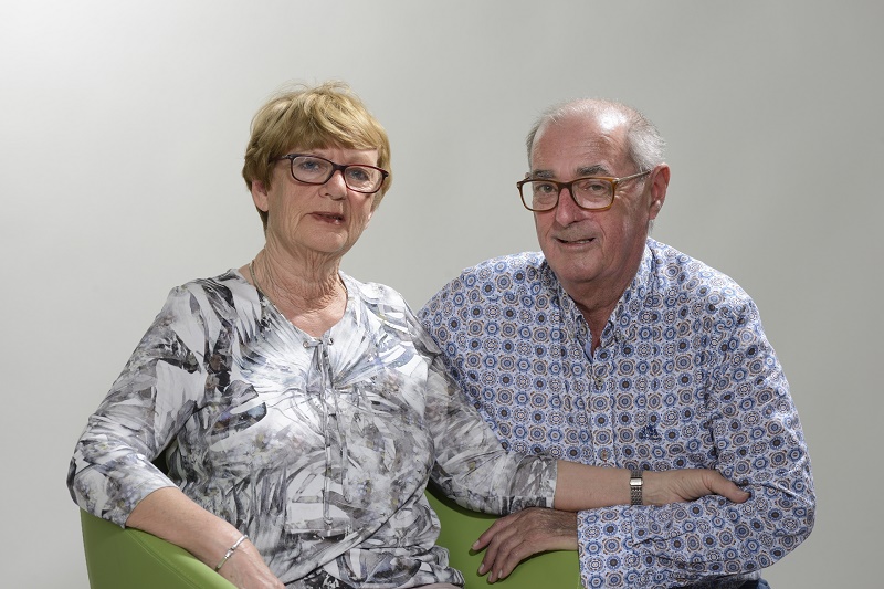 Oudere vrouw en man met brillen en geblokte shirts zitten samen voor een grijze achtergrond. Zij is dialysepatiënt.