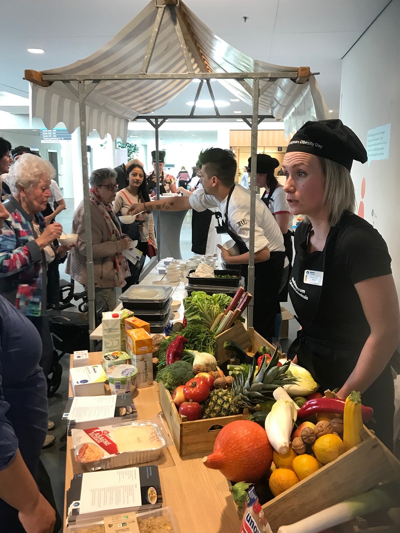 Twee mensen in kokskleding staan achter een marktkraam met verse ingrediënten en praten met een groep tijdens een etentje binnen.