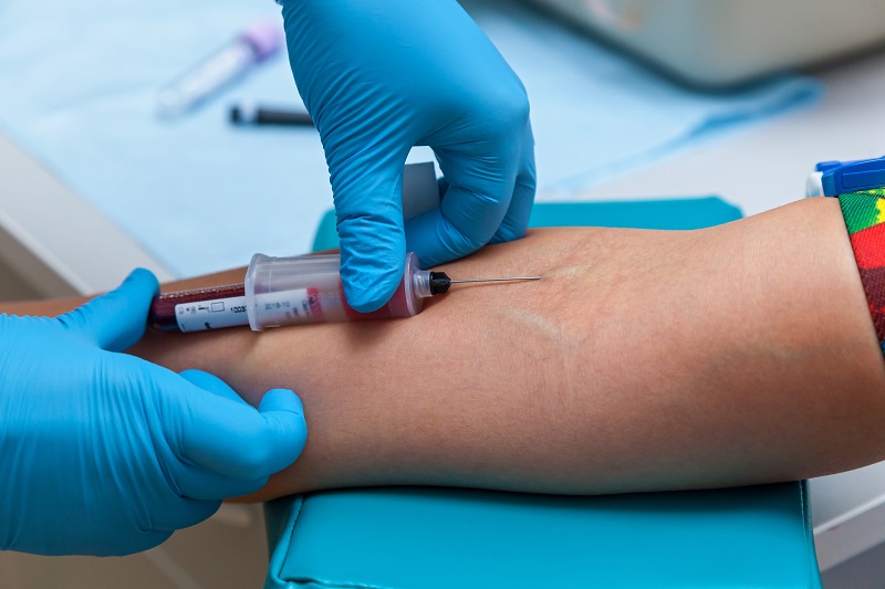 Een zorgverlener met blauwe handschoenen prikt bloed uit een arm met stuwband, de arm ligt op een steun.