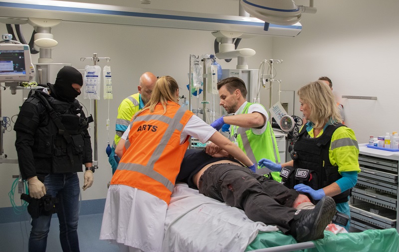 Vijf medici, waaronder één in tactische uitrusting en één met Arts-vest, verzorgen een patiënt tijdens een crisisoefening.