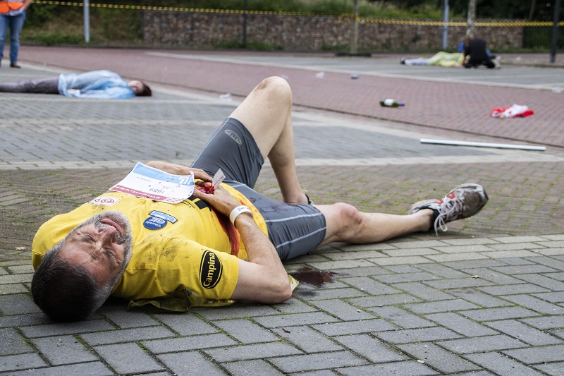 Man in geel shirt en hardloopbroek ligt pijnlijk op de grond; crisisoefening met puin en startnummers, persoon op achtergrond.