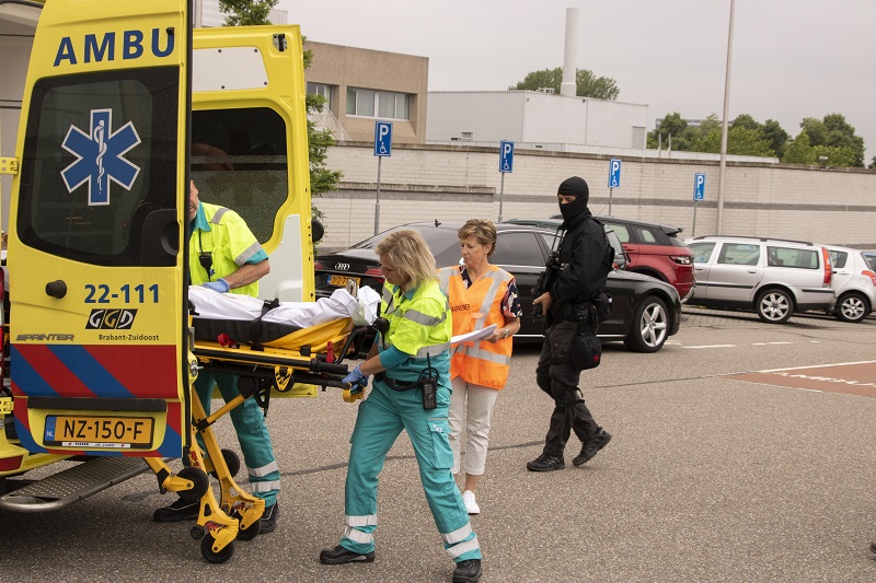 Twee ambulanceverleners laden een persoon op een brancard in tijdens een crisisoefening. Anderen lopen in de parkeergarage.