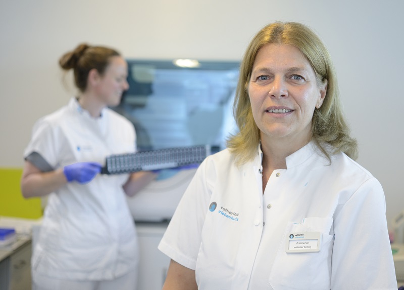 Vrouw in witte labjas lacht in laboratorium, collega op achtergrond met tray. Ze werken aan juiste behandeling en DNA-profiel.