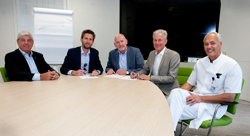 Vijf mannen, waaronder één in medisch uniform, ondertekenen samenwerking aan ronde tafel. Flipover en scherm op achtergrond.