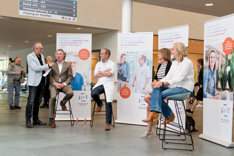 Vijf volwassenen in een paneldiscussie over nieuwe behandelingen in een ziekenhuislobby, met banners en publiek op de achtergrond.