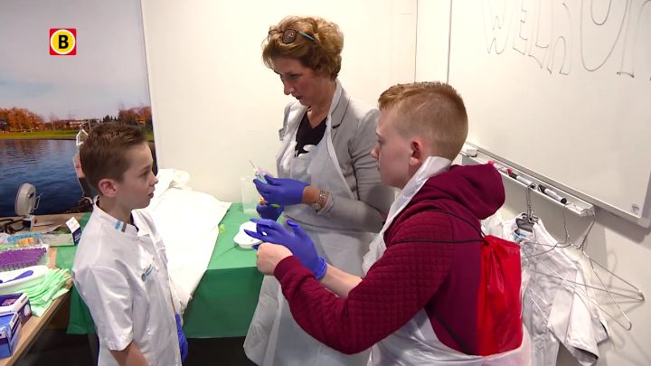 Twee kinderen met handschoenen en schorten staan met een vrouw in een labjas in een ziekenhuiskamer vol medische spullen.