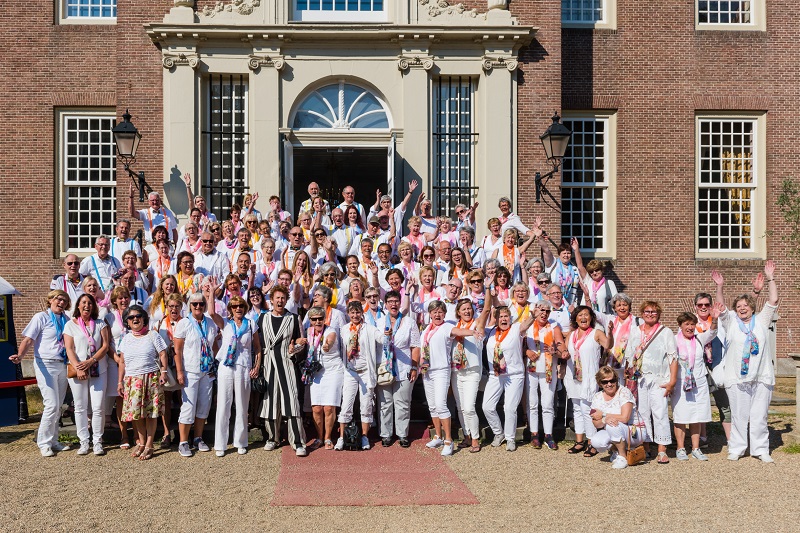 Grote groep vrijwilligers in witte kleding met gekleurde sjaals poseert lachend voor oud bakstenen gebouw in de zon.