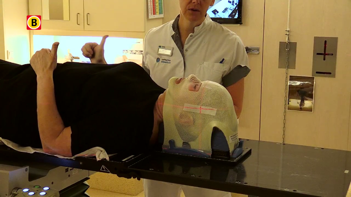 Patiënt op behandeltafel met masker op hoofd voor bestraling, zorgverlener in wit uniform erbij, medische apparatuur op achtergrond.