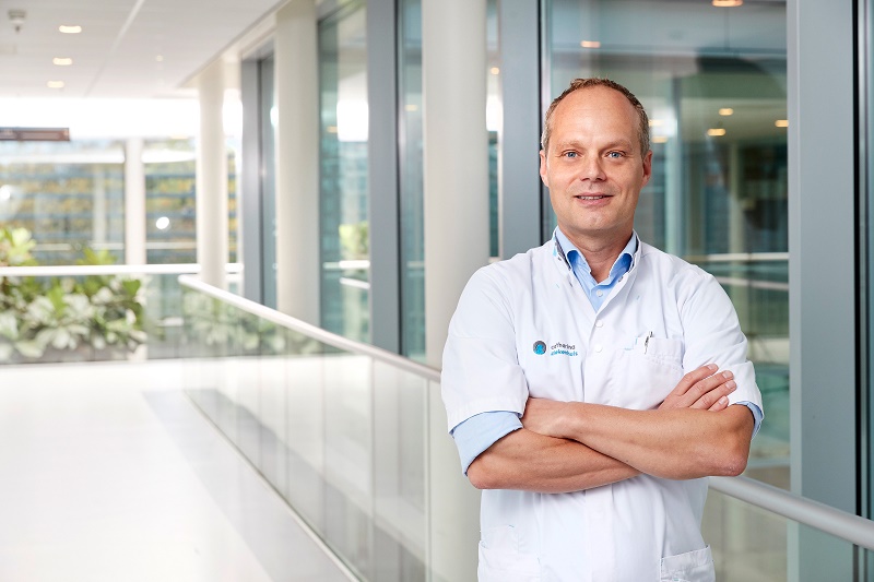Een man in een witte doktersjas staat met gekruiste armen in een lichte gang met grote ramen.