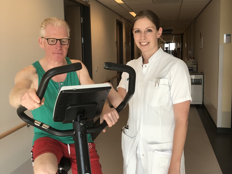 Een man met bril en groene tanktop fietst op een hometrainer in de gang, naast een vrouw in medisch uniform die lacht.