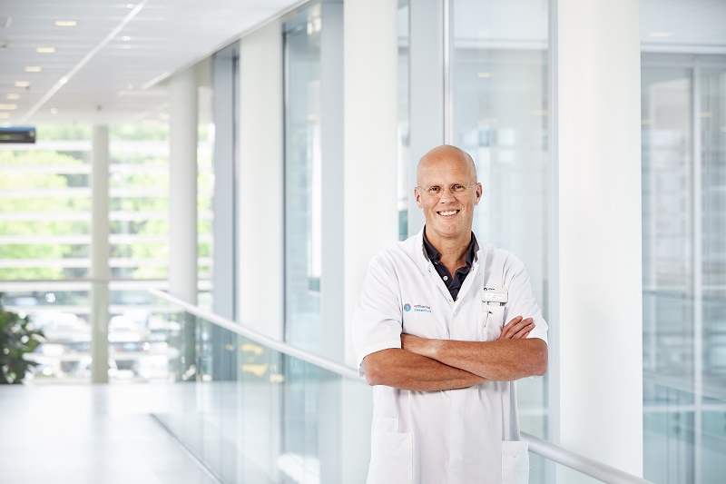 Man in witte doktersjas staat met gekruiste armen lachend in een lichte, moderne gang met glazen wanden en veel daglicht.