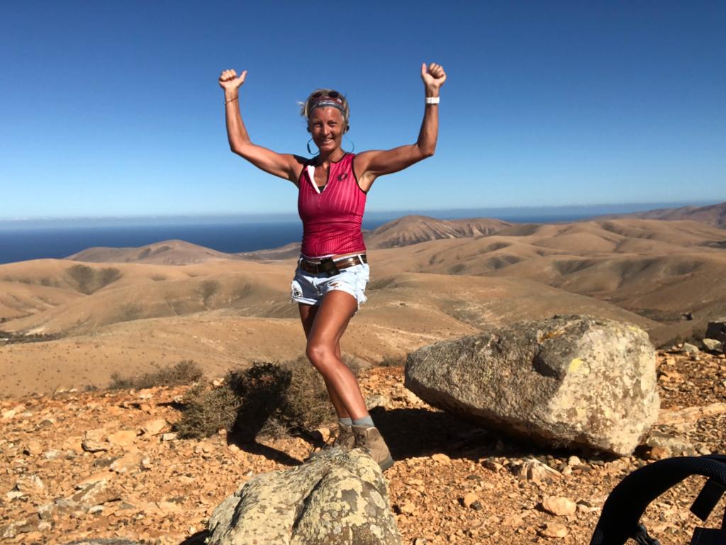 Vrouw in rode tanktop, denim shorts en wandelschoenen staat met armen omhoog op rotsen; heuvels en zee op achtergrond.