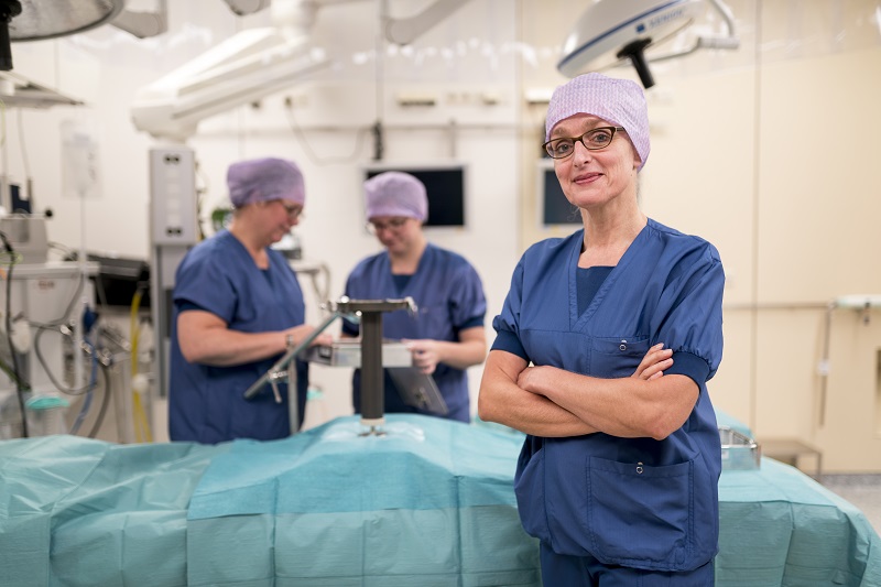 Medisch personeel in blauwe scrubs en paarse muts staat met gekruiste armen in operatiekamer; twee collega's werken op de achtergrond.