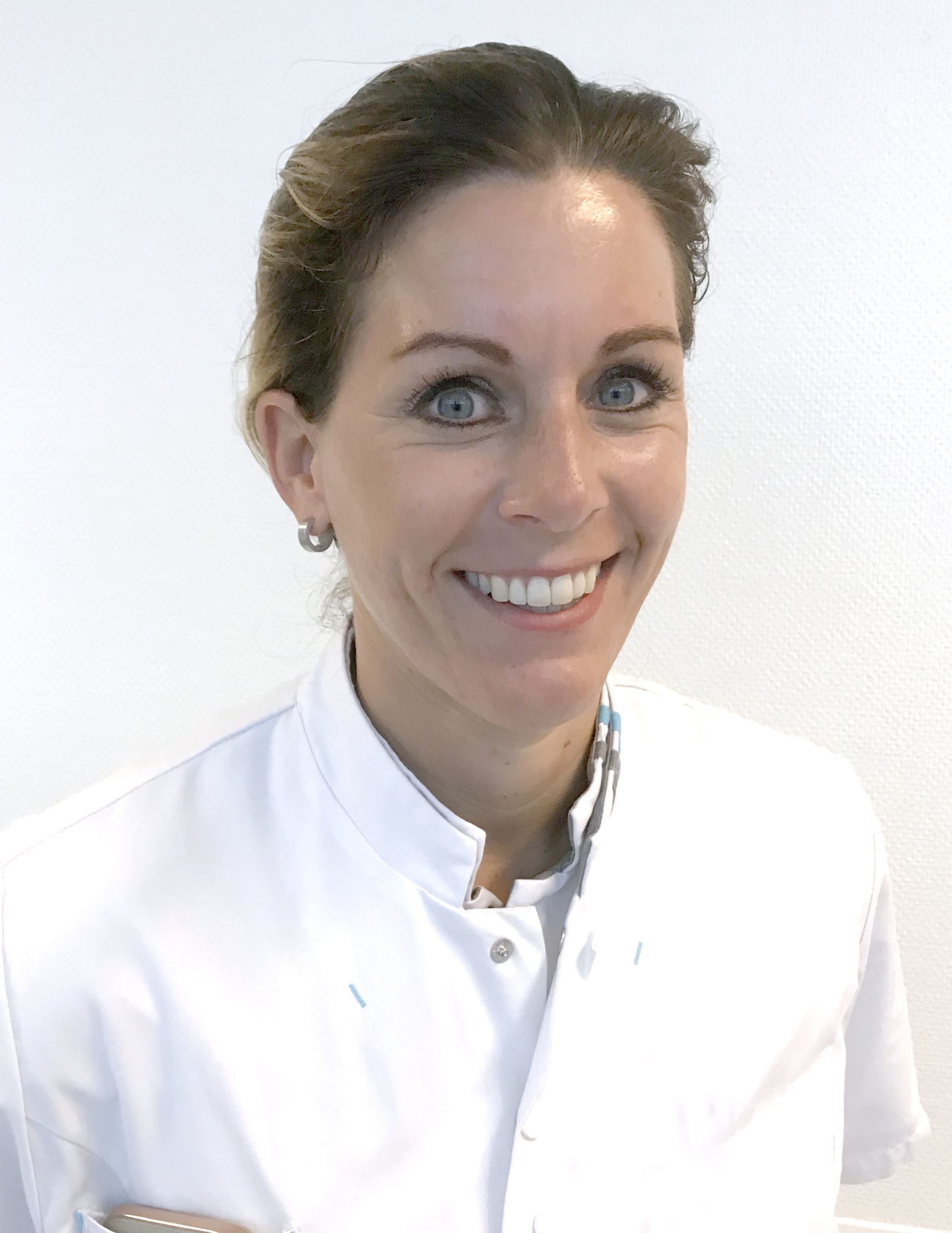 Vrouw met lichte ogen en lichtbruin haar in een witte doktersjas glimlacht naar de camera.