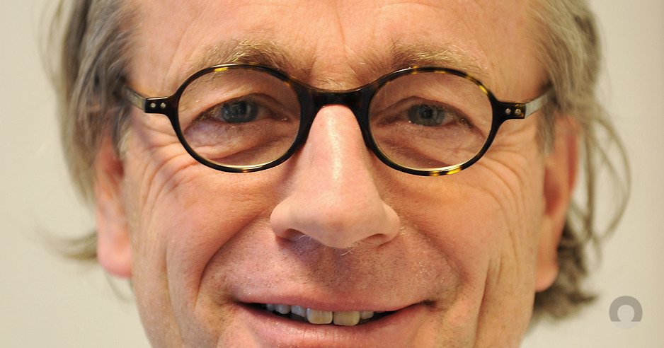 Close-up van een oudere man met grijs haar en ronde bril, lichte glimlach. Neutrale achtergrond met hint naar verbod asbestdaken.