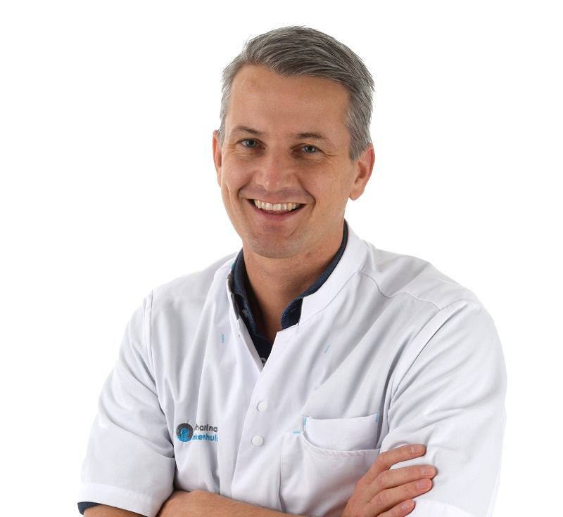 Man met kort grijs haar in witte doktersjas glimlacht met gekruiste armen, voor een witte achtergrond.