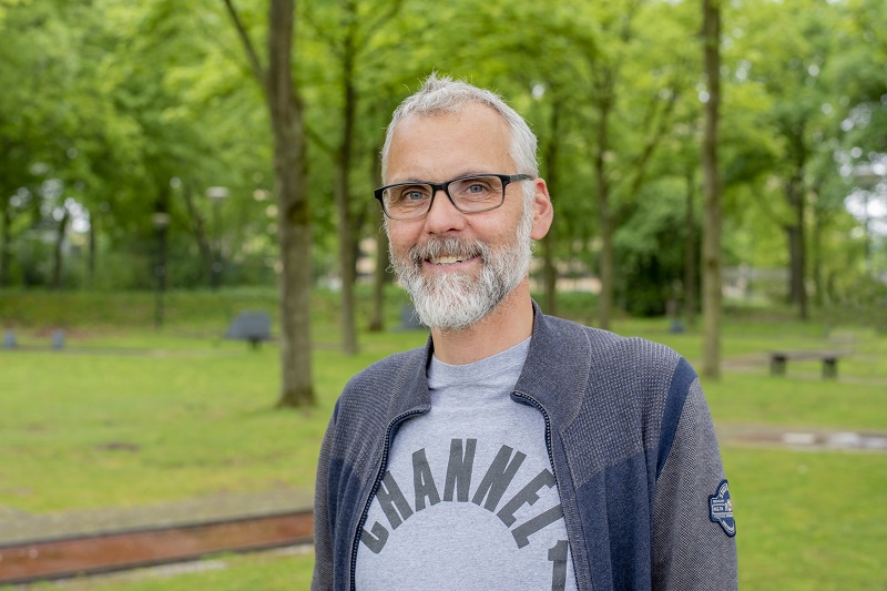 Een man met grijs haar en baard, bril, grijs shirt en donkere jas staat in een groen park en oogt normaal.