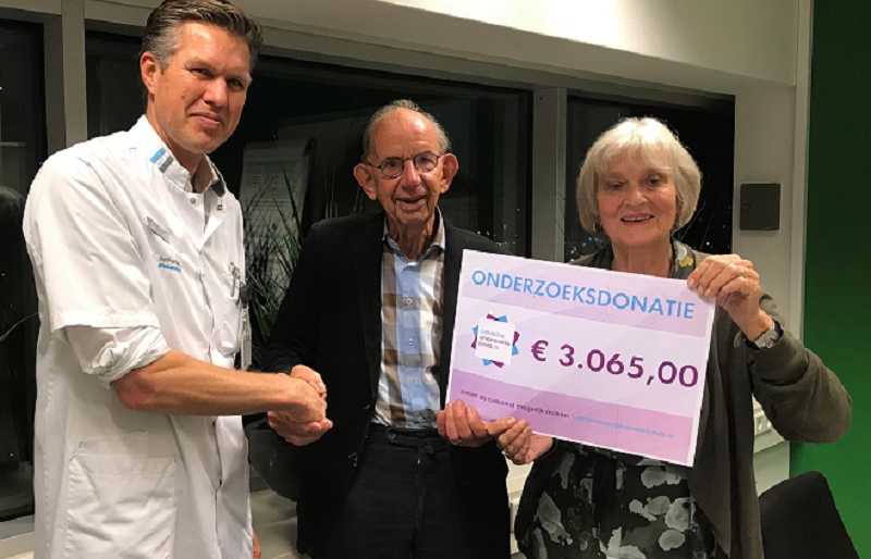 Drie mensen binnen, een kunst schudt de hand van een oudere man. Twee personen tonen een bord met 'ONDERZOEKSDONATIE €3.065,00'.