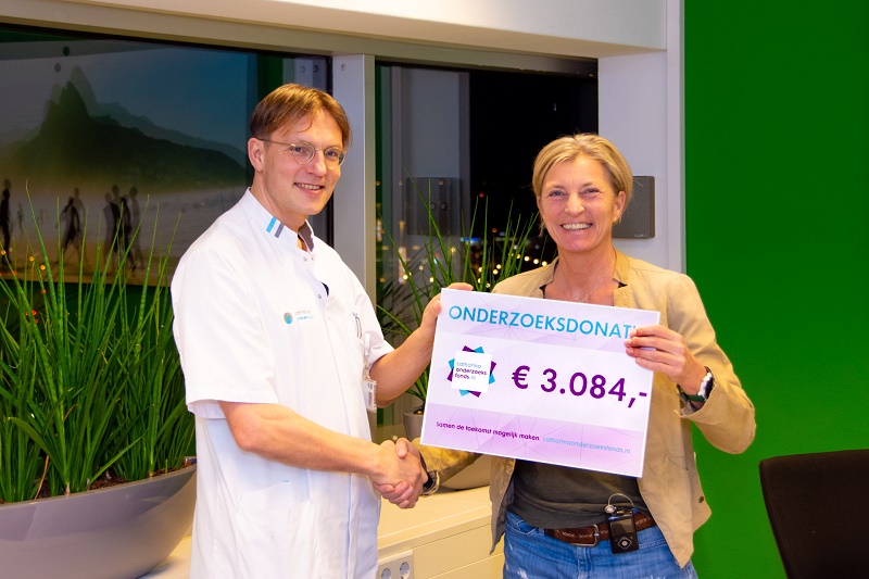 Twee mensen schudden handen en tonen een bord met "ONDERZOEKSDONATIE €3.084" naast een grote plantenbak binnen.