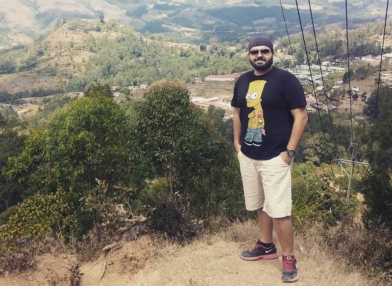 Man met zonnebril, zwart t-shirt, korte broek en rode schoenen staat op heuveltop bij herstelde vallei en bomen.