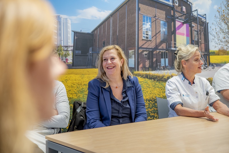 Vrouw in blauw colbert zit glimlachend aan tafel met twee mensen in witte uniformen, binnen bij foto van bakstenen gebouw.