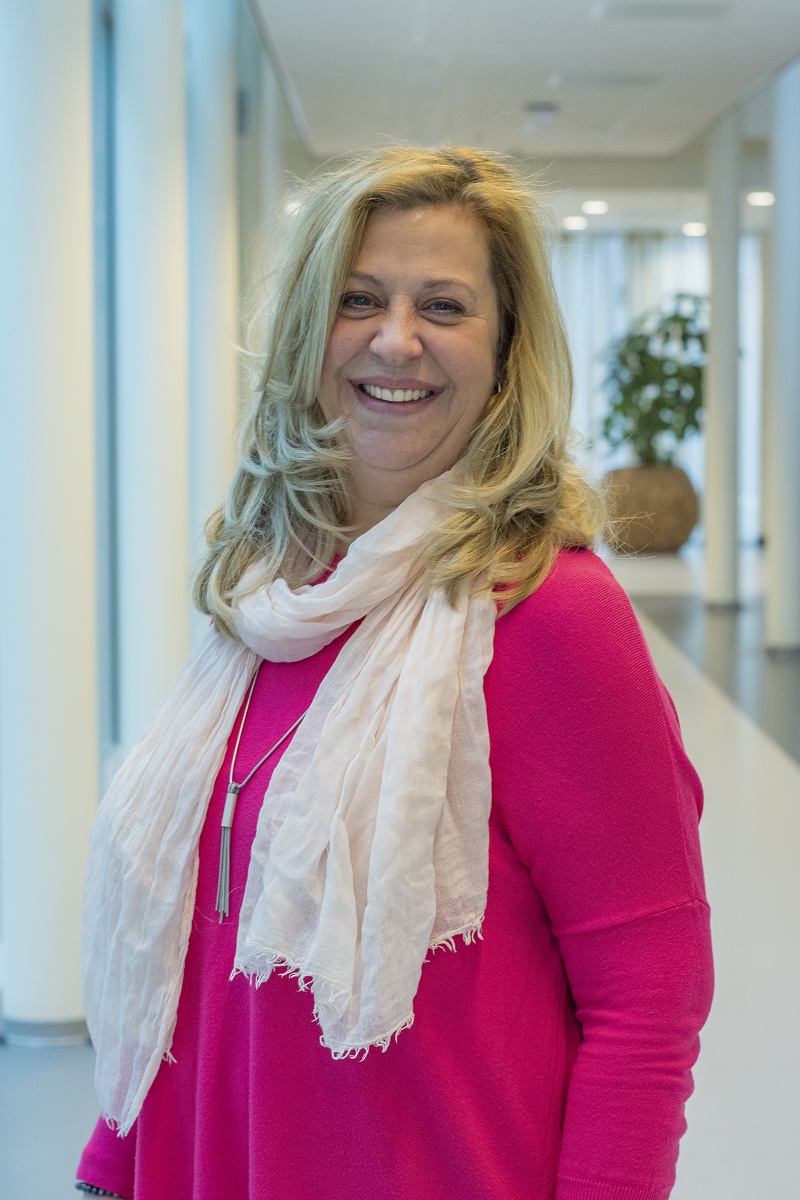 Vrouw met lang blond haar, felroze top en lichtroze sjaal lacht in moderne hal met grote ramen en witte muren.