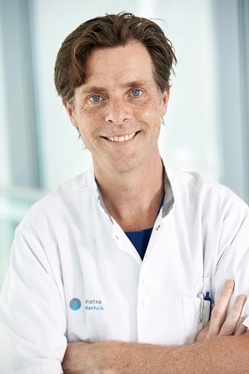 Man met bruin haar in witte doktersjas staat binnen, armen over elkaar en glimlachend. Vage blauwe achtergrond.