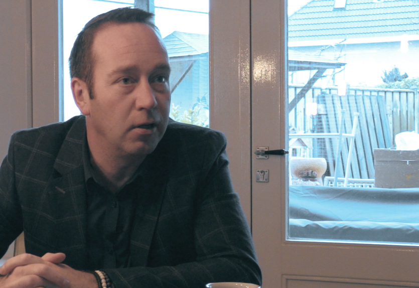 Man in geruit jasje zit aan tafel binnen, handen gevouwen. Door glazen deur tuin met mensen, tuinmeubilair en een huis zichtbaar.