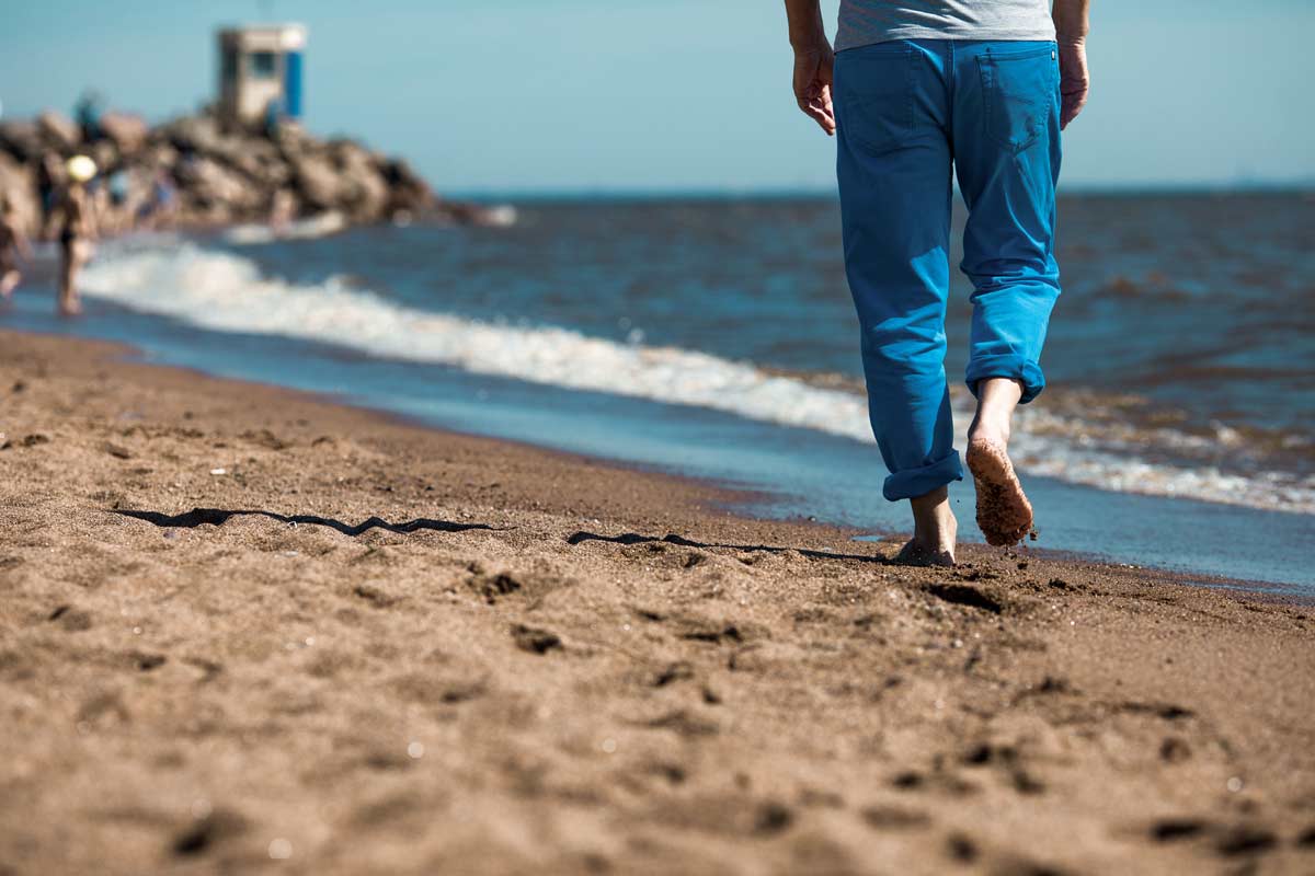 Persoon met blauwe broek loopt blootsvoets over een kalm strand; op de achtergrond enkele mensen bij zonnige structuur.