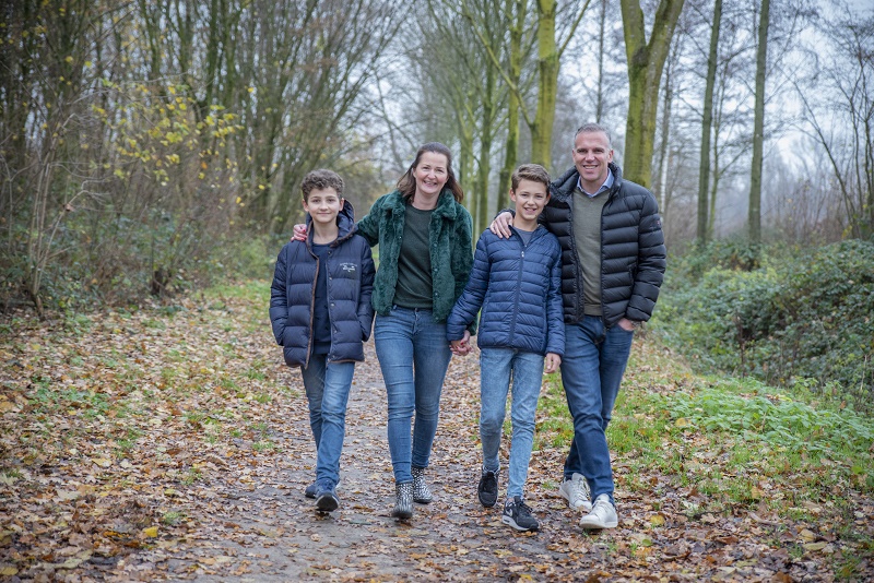 Een gezin van vier wandelt op een bladerrijk bospad. Ze dragen jassen en jeans, met bomen en boerenkool takken op de achtergrond.