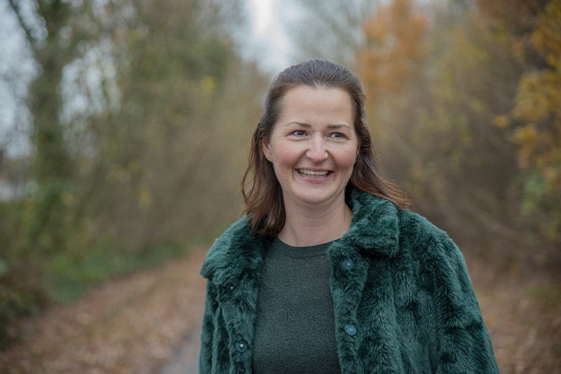 Vrouw met bruin halflang haar in groene nepbontjas staat buiten op een herfstpad en kijkt glimlachend iets naar rechts.
