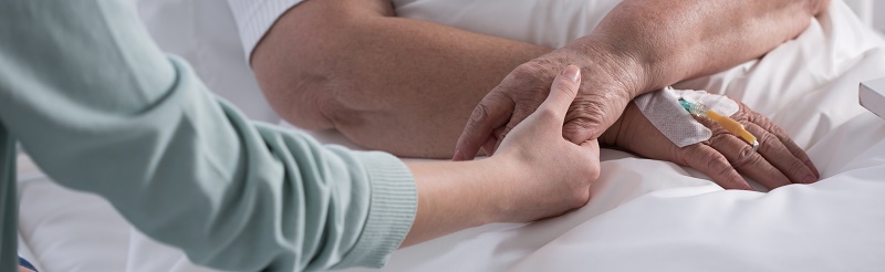 Iemand ligt in een ziekenhuisbed met een infuus en krijgt palliatieve zorg, terwijl een ander hun hand vasthoudt.