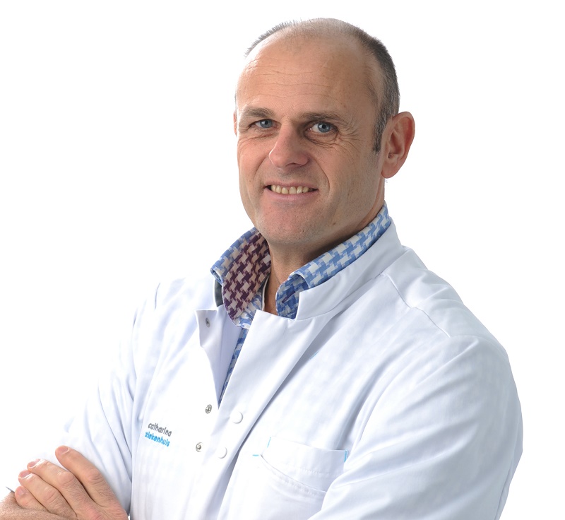 Man in witte labjas en blauw geruit overhemd met gekruiste armen voor een witte achtergrond, symbool voor preciezere radiotherapie.