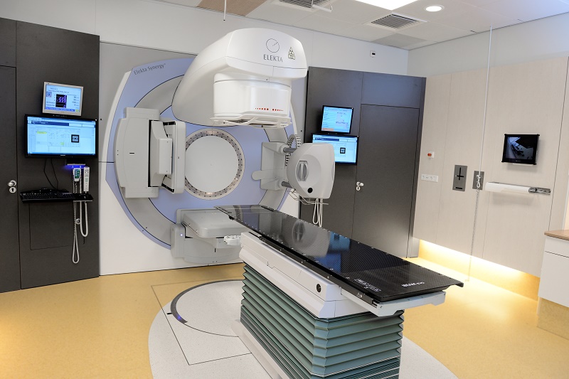 Moderne medische kamer met grote lineaire versneller, controlepanelen aan de muur en zwarte behandeltafel op gele vloer.