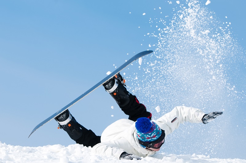 Snowboarder met witte jas, zwarte broek en blauwe muts valt voorover in de sneeuw, snowboard omhoog, sneeuw stuift op.